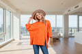 A female model wearing an orange side slit drop shoulder puff sleeve sweatshirt and a wide-brimmed hat.