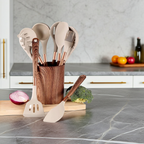 Set of kitchen utensils in a wooden holder on a kitchen counter with vegetables and fruit in the background.